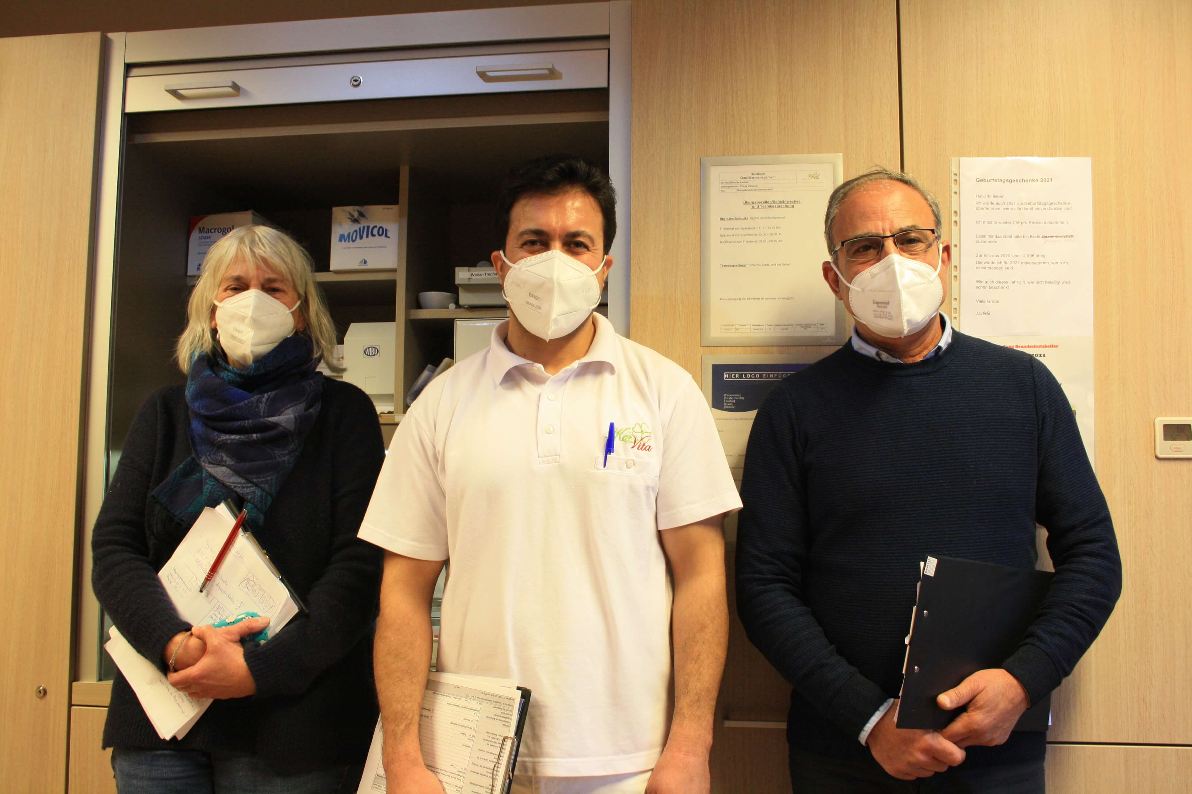 Drei Personen mit FFP2-Masken stehen nebeneinander vor einem Schrank in einem medizinischen oder pflegerischen Umfeld. Eine Frau und zwei Männer halten jeweils Unterlagen in der Hand. Die Szene vermittelt Professionalität und Hygienebewusstsein im Gesundh